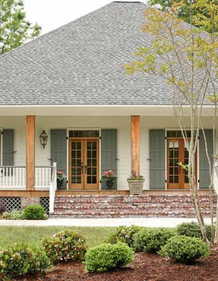 Design by Krista Lewis, K. Lewis Design.  Photography by Nancy Nolan for At Home in Arkansas. Acadian cottage exterior Brick Southern Home Exterior, White House With Natural Wood Shutters, Olive Green And Cream House Exterior, Trendy Shutter Colors, Exterior Shutters Ideas Brick, White House With Stained Shutters, Covered Porch Paint Colors, Exterior Oval Window, White And Green Home Exterior