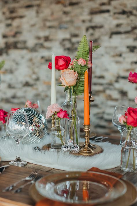 We had so much fun styling this boho disco themed shoot! We used vintage elements in combination with classic boho decor to make a super fun and unique table. These long farm tables are great for styling unique centerpieces. For ours, we used a combination of vintage candlestick holders, bud vases filled with assorted pink flowers, shiny disco balls, and went with a fun fur throw as a table runner. Sparkly and metallic gold chargers looked just right with a warm brown napkin. Shot by @kaboovang Disco Ball Wedding Tables, Wedding Table Decor Disco Balls, Fun Flower Vases, Centerpieces With Disco Balls, Pink Bud Vases Wedding, Disco Centerpieces Wedding, Disco Wedding Flowers, Disco Ball Wedding Table Decor, Disco Wedding Table Setting