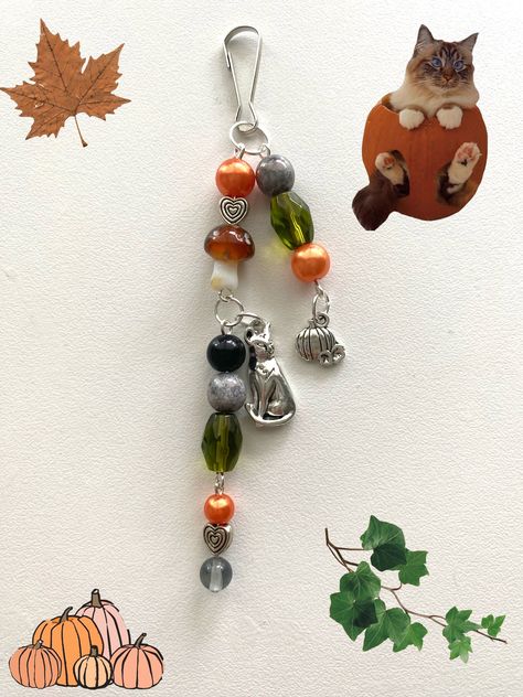 A beaded keychain with two silver charms. The pumpkin and cat charms along with the orange, green, and gray beads give off the fall aesthetic. The keychain also includes a glass mushroom bead, adding to the overall fall appearance. Fall Keychain Ideas, Keychain Ideas Diy Beads, Halloween Beaded Keychains, Beaded Keychains Aesthetic, Diy Charms Keychain, Keychain Beads Ideas, Fall Keychains, Beaded Keychain Ideas, Keychains Aesthetic