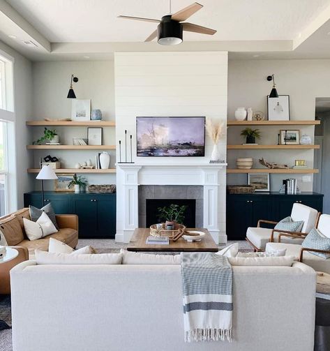 This modern living room is designed with a white fireplace and traditional mantle, which is accessorized with a vase of dried pampas grass, candlesticks, and painting. Warm teal cabinets are built on either side, above which hang light wood floating shelves. Light Wood Floating Shelves, Floating Shelves Fireplace, Fireplace With Cabinets, Shelves Around Fireplace, Shelves Fireplace, Built In Around Fireplace, High Ceiling Living Room, Floating Shelves Living Room, Shelf Decor Living Room