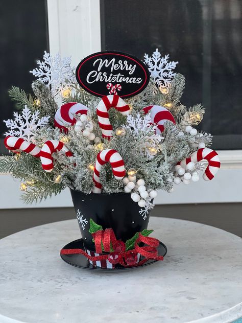 Celebrate the joy and wonder of the winter season with this enchanting snowman hat centerpiece. This unique piece is the epitome of holiday cheer, featuring a charming snowman hat surrounded by festive candy canes, twinkling lights, and lush winter pines. Whether it's the centerpiece of your holiday table or a cheerful addition to your mantel, this delightful arrangement is sure to bring smiles and a cozy, festive atmosphere to your home. Details: *Composition includes a decorative snowman hat, artificial candy canes, LED string lights, and faux winter pine branches *Battery-operated lights for easy and safe use (batteries not included) *Perfect for holiday tablescapes, mantel decor, or as a festive accent piece Snowman Table Setting, Mason Jar Winter Centerpieces, Snowman Table Decorations Centerpieces, Candy Cane Centerpiece Ideas, Snowman Centerpiece Ideas, Christmas Centerpieces Diy Table, Snowman Hat Centerpiece, Holiday Centerpieces For Tables, Diy Christmas Hats