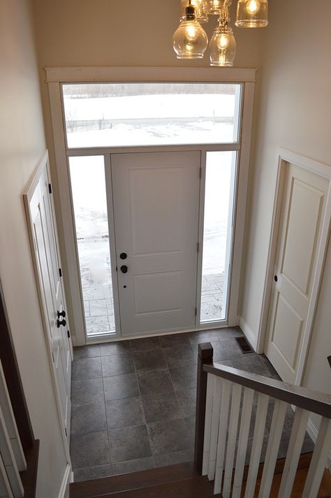 Front entry with grey tile and hardwood stairs. Raised bungalow front hall. Transom window above front door. Jar lights. Front Door With Transom Above, Double Front Door With Transom Above, Mudroom With Transom Window, Window Above Front Door, Transitional Kitchen Backsplash, Front Hall Decor, Single Front Door With Sidelights And Transom, Single Door With Transom, Raised Bungalow