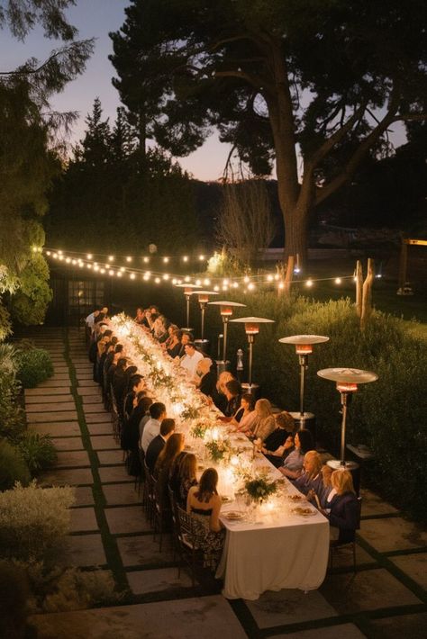 A long table set for an elegant outdoor dinner is surrounded by guests seated under string lights. The evening sky is dusky, and trees create a natural backdrop. Warm lighting and greenery add to the intimate ambiance. Feast Table Wedding, One Long Wedding Table, Wedding Table Long Layout, Two Long Tables Wedding, Backyard Wedding Long Table, Wedding Table Configuration, Outdoor Long Table Dinner, U Shaped Wedding Reception, Long Banquet Tables Wedding