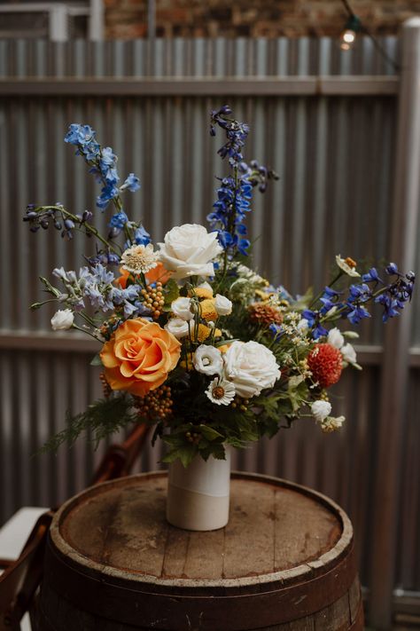 Romee Willow Florals, an artful florist in Chicago, created this elegant, organic, colorful, floral wedding ceremony arrangement. Flowers used were blue delphinium, orange roses, white roses, orange dahlias, yellow ranunculus, and white zinnias. This late summer wedding took place at Firehouse Chicago in August. Photo by Mark Trela Photography. To see more work by Romee Willow Florals, click the link. Red Yellow Blue Wedding Flowers, White Orange Blue Wedding Bouquet, Event Flowers Arrangements, Simple Colorful Floral Centerpieces, Blue Yellow Orange Flower Arrangements, Blue And Orange Flowers Wedding, August Wedding Flowers Centerpieces, Blue White Orange Flower Arrangements, Orange Blue Wedding Flowers