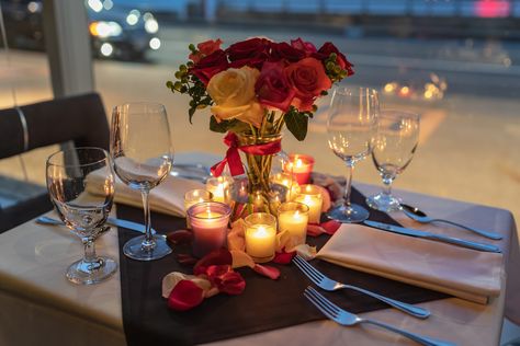 A restaurant table decorated with a vase of red, pink, and white flowers. The vase is surrounded with candles and tableware. Proposal Dinner Table Settings, Romantic Dinner Table Setting For Two, Romantic Dinner Table Setting, Rose Petals And Candles, Proposal Dinner, Bride Proposal, Romantic Dinner Tables, Engagement Proposals, Engagement Plan