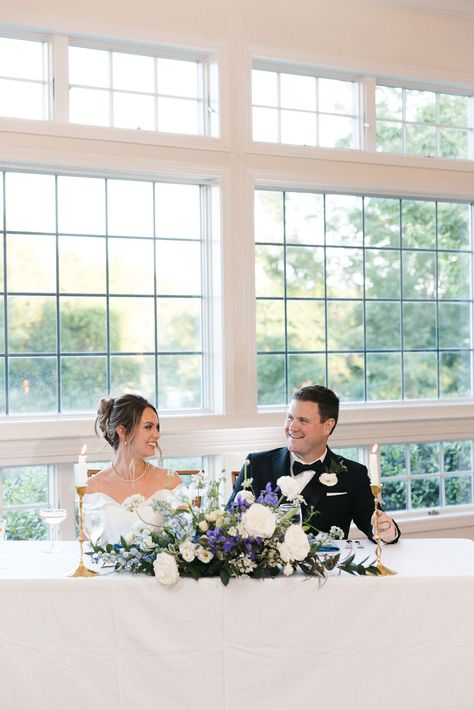 Image of a bride and groom at their sweetheart table at their reception at the Coonamessett. The table features a white, blue and green statement floral design with white peonies, ranunculus, greenery and more. Blue And White Sweetheart Table, Sweetheart Table Blue, Blue Sweetheart Table, White Sweetheart Table, Sweethearts Table, Coastal White, Sweetheart Table Wedding, Coastal Elegance, Table Wedding