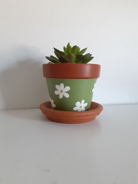 A green terracotta pot with white flowers; also comes with the saucer. Has a small hole in the bottom for drainage. Great for small plants such as succulents or a small house plant! Flowers Pot Painting, Painting Succulent Pots, Small Terra Cotta Pot Ideas, Small Plants Aesthetic, Cute Pots Painting Ideas, Painting Ideas On Plant Pots, Simple Pot Designs, Small Terracotta Pot Painting Ideas, Hand Painted Terracotta Pots