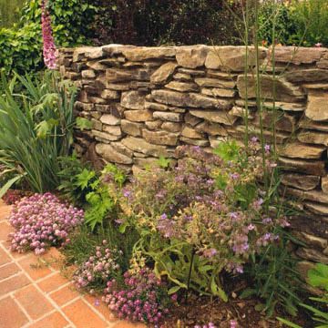 A dry-stacked stone wall looks so natural. The spaces between stones in the retaining wall allow water to drain through, relieving pressure. Another advantage of dry-stacked stone walls is that you can create planting pockets for small rock garden plants such as sedums. Garden Landscaping Design Ideas, Building A Stone Wall, Stone Walls Garden, Dry Stack Stone, Stacked Stone Walls, Gravel Landscaping, Backyard Garden Layout, Rock Garden Plants, Rock Garden Design