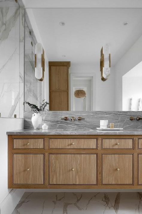 Antique brass faucets are fixed to a gray marble slab backsplash under a frameless vanity mirror fitted with brass and alabaster oblong sconces. Angled Cabinet, Transitional Guest Bathroom, Marble Slab Backsplash, Slab Backsplash, Madison Ct, Antique Brass Faucet, Marble Bathroom Floor, Brass Faucets, Marble Wall Tiles