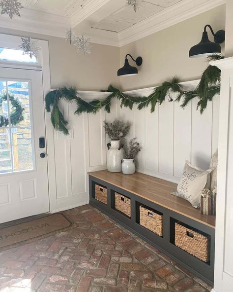 This entryway features cream-colored walls, white wainscoting, and a red brick herringbone floor. Branches in white jugs are placed on a long entryway bench with storage. Two black sconces illuminate the garland draped over the white wainscoting. Entryway Ideas Long Entry Hall, Entryway Wainscoting Ideas, Wainscoting Ideas Entryway, Long Entryway Bench, Entryway Shiplap, Laundry Room Wall Paint, Brick Entryway, Wainscoting Entryway, Brick Herringbone Floor