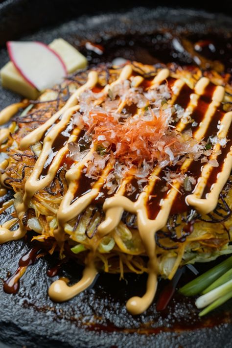 Okonomiyaki topped with drizzles of mayonnaise, a savory sauce, and bonito flakes on a dark plate, garnished with sliced radish. Japanese Okonomiyaki Recipe, Japanese Pancake Okonomiyaki, Traditional Japanese Dinner, Oyaki Japanese, Traditional Japanese Food Recipes, Yaki Imo, Sukiyaki Recipe, Eggplant Rollatini Recipe, Okonomiyaki Recipe