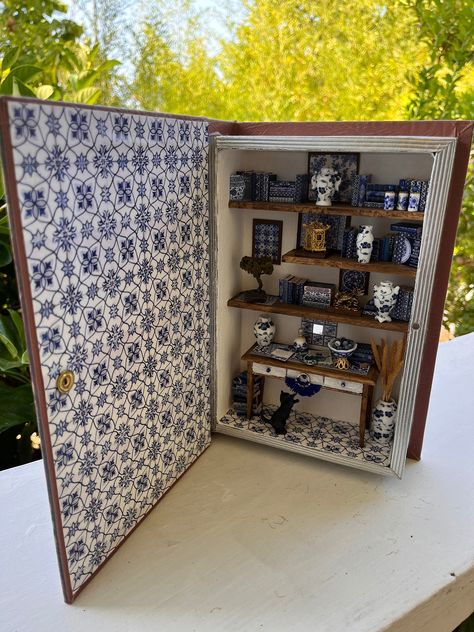 Beautiful miniature bookcase inserted in a book-shaped box, all themed around white and blue oriental ceramics! In this beautiful book (it is a reproduction, a box made in the shape of a book with a white and blue cover) there is a bookcase themed with white and blue oriental majolica and the oriental theme is repeated through various objects inserted in the setting: a small bonsai , a small golden pagoda, a paperweight made in the shape of a dragon and other small elements All glued as in the photo and carefully studied down to the smallest detail! A kitten plays with the blue tassel hanging from the desk drawer knob, bringing the scene to life! unique piece, all in 1:12 scale Faux Book Storage Boxes, Miniature Scenes Diy, Book Diarama Ideas, Book Box Ideas, Small Book Case, Mini Bookcase, Miniature Bookcase, Cartonnage Boxes, Book Miniature