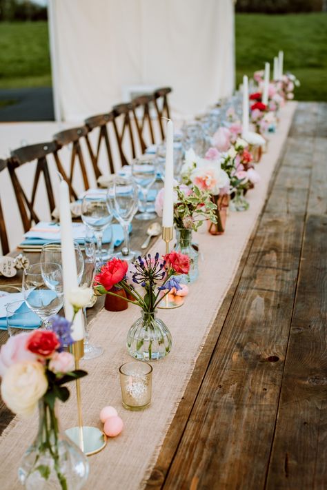 Pops of colour in low bud vases worked really well with the hessian runner and natural wooden tables for this Easter wedding reception in a clear span marquee at Barnsley House in the Cotswolds, UK. We add tealight votives in mercury gold glass, slim dinner candles and miniature chocolate eggs to complete the styling. Spring Wedding Long Table Decor, Wedding Tables With Bud Vases, Long Table Centrepiece Wedding, Long Wooden Wedding Table Decorations, Flowers Candles Centerpiece, Wedding Table Decorations Wooden Table, Mix And Match Wedding Tables, Rustic Wedding Table Flowers, Wedding Table Tealights