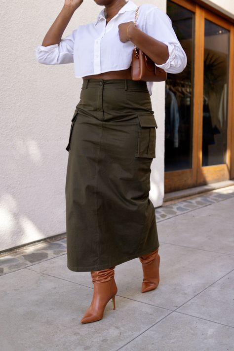 Black woman wearing an olive green cargo maxi skirt with cognac colored slouch boots, a white cropped button up, and a cognac colored purse