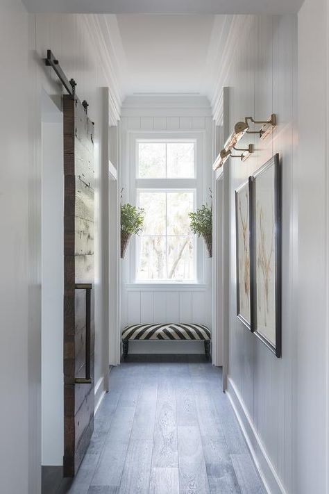 Chic hallway features vertical shiplap walls lined with side by side art illuminated by antique brass picture lights situated across from a plank barb door on rails. Narrow Hallway Lighting, Palmetto Bluff Homes, End Of Hallway, Hallway Art, Upstairs Hallway, Hal Decor, Hallway Designs, Hallway Design, Long Hallway