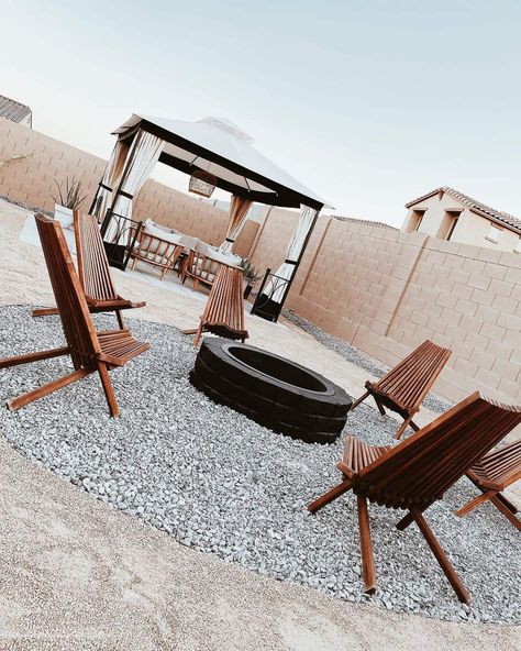 A modern patio has a circle of gray gravel, and a black stone fire pit resting inside. Wooden patio chairs with clean lines are arranged around the fire pit. In the background, a covered pergola is arranged over the top of wooden patio furniture. Wooden Patio Chairs, Wooden Patio Furniture, Wooden Patio, Brick Fire Pit, Fire Pit Chairs, Metal Fire Pit, Wood Fire Pit, Cozy Backyard, Stone Fire Pit