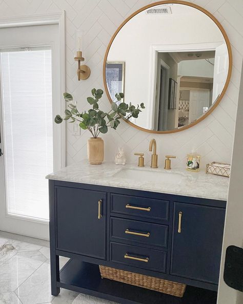 A round gold mirror hangs on a white herringbone tile bathroom wall in this modern bathroom. Gold hardware is installed on the navy blue cabinet on a gray tile floor. Place Easter dÃ©cor, such as a white rabbit next to a wood vase filled with green branches, for a bright, Springtime touch. White Herringbone Tile Bathroom Wall, Bathroom Gold Hardware, White Herringbone Tile Bathroom, Gray Tile Floor, Navy Blue Cabinet, Navy Blue Vanity, Herringbone Tile Bathroom, Blue White Bathrooms, White Herringbone Tile