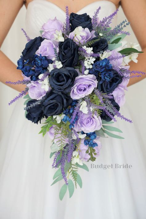 This lovely assortment of navy blue and lavender roses is bound to turn heads on your wedding day. This lovely cascading brides’ bouquet from the is an absolutely breathtaking arrangement for any bride to be, in any season. Sage greenery and tons of lavender fronds complete the look and add a delicate realism to this beautiful bouquet. This bouquet is 10” wide, 18” long, and is crafted by our in-house florists without using a foam base to anchor the flowers to the bouquet. Instead, we wire our b Navy And Wisteria Wedding, Navy Blue Lavender Wedding, Navy Blue And Lavender Wedding Ideas, Plum And Navy Blue Wedding, Navy Blue And Purple Wedding Flowers, Blue And Purple Bouquet Wedding, Navy Blue And Lilac Wedding Theme, Purple And Navy Wedding Flowers, Navy And Lavender Wedding Flowers