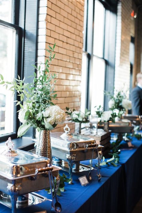 One of the last places couples think to decorate is the buffet table, but sprucing up this space will add some wow factor to your wedding. This couple used a navy linen instead of the typical black, and included some simple vases with a white hydrangea and greenery. Simple, classy, and sure to impress guests. Buffet Dishes Display, Chafing Dish Display Ideas Wedding, Buffet Wedding Decor, Party Buffet Table Set Up, Rehearsal Dinner Buffet Table Decor, Buffet Centerpiece Ideas, Elegant Buffet Table Ideas Decor, Elegant Wedding Buffet Ideas, Wedding Buffet Tables