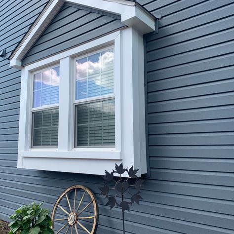 The photo is a close up on a home showing it's greyish-blue vinyl siding with large center window that has white trim. There's also a wagon wheel as decor against the home, and a large green plant, along with a windmill. In the reflection of the window, you can see the clear sky and fluffy clouds. Can You Paint Vinyl Siding On A House, Painted Vinyl Siding House Exteriors, How To Paint Vinyl Siding On A House, Outside Vinyl Siding Colors, Can You Paint Siding On A House, Updating Vinyl Siding Exterior, Can Vinyl Siding Be Painted, Painting Vinyl Siding House Exteriors, Painting Exterior Vinyl Siding