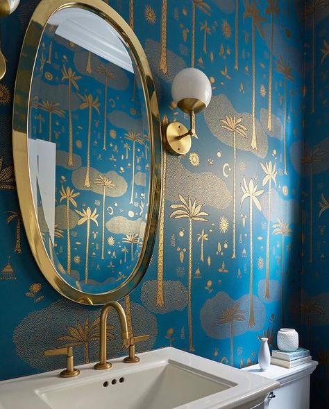 Striking blue and gold wallpaper accents an elegant contemporary powder room holding a gold oval mirror between gold and white glass globe sconces mounted over a white pedestal sink finished with a brushed gold faucet. Blue And Gold Wallpaper, Contemporary Powder Room, Powder Room Wallpaper, Gold Faucet, Tropical Bathroom, Teal Wallpaper, Gold Wallpaper, Bathroom Wallpaper, Elegant Interiors