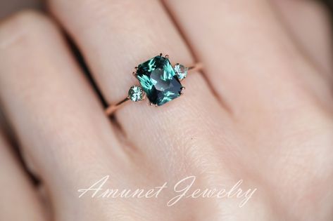 This ring features Stunning deep green cushion sapphire in a cluster setting enhanced by two teal green Montana sapphire on the side.Main ring specification:Main stone: Natural teal Madagascar sapphire (unheated)Dimension: 7.83mm x 6.14 mWeight: 2.1 CtClarity: VSShape: CushionSide stones: Montana sapphire 2.5mm(2).Material: 14K yellow, rose or white goldAvailable Sizes: USA 3 - USA 9 (larger & smaller sizes available. Price on request)At the time of selecting the ring please choose your band col Sparkly Rings, Sapphire Ring Engagement, Teal Sapphire Ring, Colored Stone Engagement Rings, Green Montana, Green Sapphire Ring, Sapphire Wedding Band, Sparkly Ring, Blue Green Sapphires