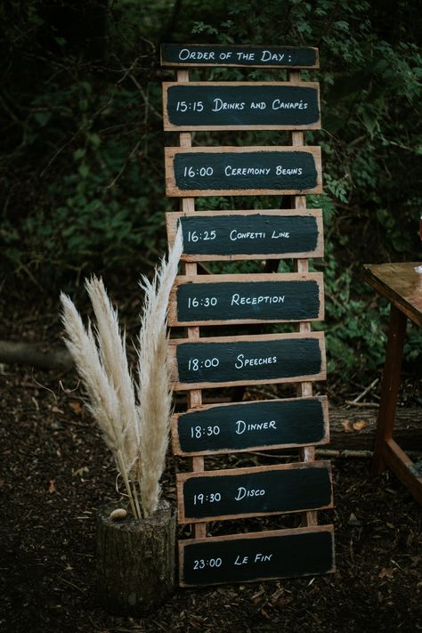 This shows an outdoor wedding. The focus is on a 'order of the day' sign in a rustic wooden black board sign. Next to it are some dried pampas leaves displayed on a trunk of wood. Order Of Service Wedding Sign, Blackboard Sign, Order Of The Day Sign, Dyi Wedding, Wedding Guest Signing, Lloyd Jones, Wood Photography, Spa Ideas, Plan A Wedding