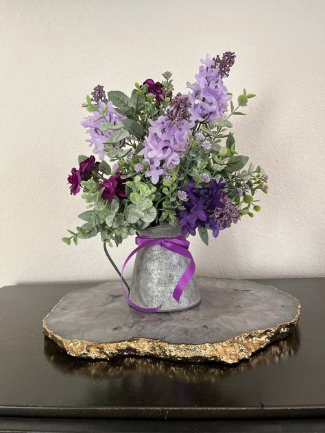 Transform your space into floral paradise with this exquisite roses and lilac flower arrangement in stunning shades of purple and lavender. This beautiful centerpiece features a blend of artificial miniature roses, lilacs, and eucalyptus for that touch of elegance. You will find your arrangement in a charming galvanized watering can or wash tub with wooden handles that adds that rustic country flair to any decor. Then it is topped with the finishing touch of a beautiful tied purple bow that will Simple Purple Table Decorations, Small Purple Centerpieces, Purple Floral Party Decor, Purple And Green Wedding Centerpieces, Purple Centerpiece Ideas, Lavender And Sage Wedding Decorations, Lilac Table Decor, Lilac Centerpieces, Table Decorations Purple