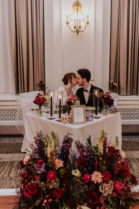 Dark holiday vibes abounded at Celia + Pablo’s “eve of the eve” December 30 wedding. Deep burgundy reds, rich emerald greens, and eggplant purples combined to create a romantic atmosphere. Celia’s bouquet included red garden roses, blue anenomes, purple ranunculus, raspberry stock, and red orchids. Pablo’s boutonniere featured a red orchid with seasonal greenery and berries. Antique gold compotes overflowing with flowers made their  author-themed guest tables gorgeous places to gather. Deep Red And Green Wedding, Deep Red And Purple Wedding, Purple And Red Wedding, Head Table Flowers, Moody Wedding Flowers, Purple Ranunculus, Red Orchid, Berry Wedding, Head Table Wedding