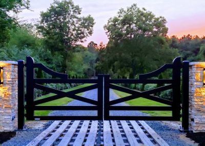 The sunset makes this farmhouse-style gate entrance picture perfect! These aluminum gates were faux-wood painted to add detail and give them texture. Check out more photos and details on this gate at this link. We'd love to help you with your dream gate! Farmhouse Driveway Entrance, Farm Gates Entrance, Ranch Entrance Ideas, Entrance Gates Driveway, Aluminum Gates, Property Gates, Gated Entrance, Metal Driveway Gates, Driveway Entrance Landscaping