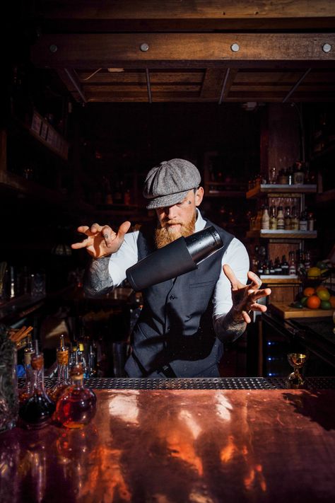 Bartender with beard and vintage hat flipping cocktail shaker mid air at the bar with bottles and wooden shelves around him Bartenders Photography, Boston Style, Bartender Tools, Most Popular Cocktails, Tiki Mugs, Popular Cocktails, Boston Fashion, Restaurant Photography, Cocktail Shaker Set