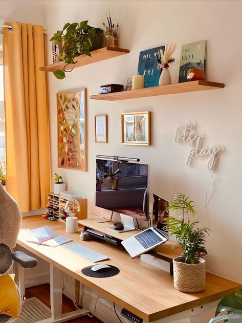 Cozy home office setup featuring a wooden desk under sunlight from a window with yellow curtains. The space is decorated with green plants on floating shelves, motivational posters, and a neon sign saying 'let's play' above the desk. A laptop and monitor provide a functional work area. Cozy Desk Setup, Cozy Workspace, Interior Design Layout, Cozy Desk, Work Office Decor, Dorm Room Designs, Dream Apartment Decor, Desk Inspiration, Ideas Para Organizar