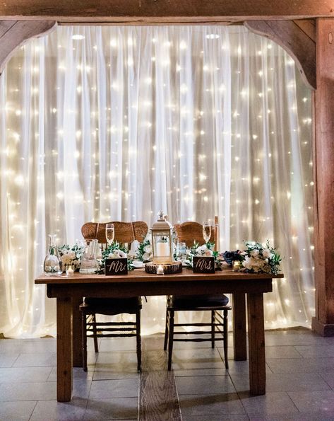 Sheer curtain overlaying lights for a beautiful backdrop behind the sweetheart table Wedding Signs Behind Head Table, Bride And Groom Table Lights, Curtain Lights Wedding Tent, Backdrop Curtain Lights, Sweetheart Table Curtain Backdrop, Curtain Light Backdrop Wedding, Behind Wedding Table Decor, Sheer Curtain Backdrop With Lights, Fairy Light Wall Wedding