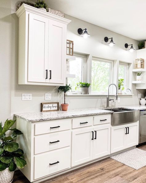 These white cabinets with black hardware are topped by a gray and black countertop in this farmhouse kitchen. A stainless apron sink is fitted into the counter under white windows. There is no backsplash behind the sink. Kitchen Ideas Marble Countertops, White Dream Kitchen, Over Kitchen Sink Lighting, Kitchen Sink Lighting Ideas, White Tile Kitchen Backsplash, Kitchen Sink Lighting, White Tile Backsplash, Organizing Hacks, Farmhouse Kitchen Design