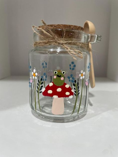 Hand painted frog mushroom jar and spoon with wildflowers all the way round. Would make a great unique gift or something different to add to your own collection ! 🍄🐸 250ml capacity  Please note : glasses are hand painted and so designs may differ ever so slightly from product image  Hand Wash only  Do not soak ! Pencil Holder Painting Ideas, Hand Painted Jars, Glass Jar Painting Ideas, Mason Jar Painting Ideas, Painting Jars, Painted Glass Jars, Glass Jars Diy, Painted Frog, Mushroom Jar