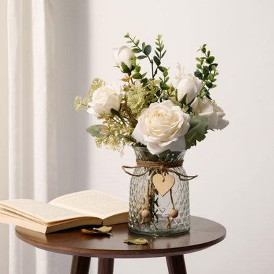 Love blossoms in details. This artificial flower bouquet paired with a glass vase adorned with a heart-shaped pendant, whispers tenderness. The elegance of white roses and freshness of eucalyptus create a cozy ambiance. As a table centerpiece or room accent, it lets love, like this exquisite bouquet, bloom quietly in every corner. | Primrue Faux Flower Arrangement w/ Vase, Silk | C111485239 | Wayfair Canada Small White Rose Centerpiece, Wedding Flower Vases, Wedding Vases Centerpieces, Fake Flowers Bouquet, Sage Christmas, Flowers With Vase, Eucalyptus Flowers, Sage Green Baby Shower, Rose Centerpiece