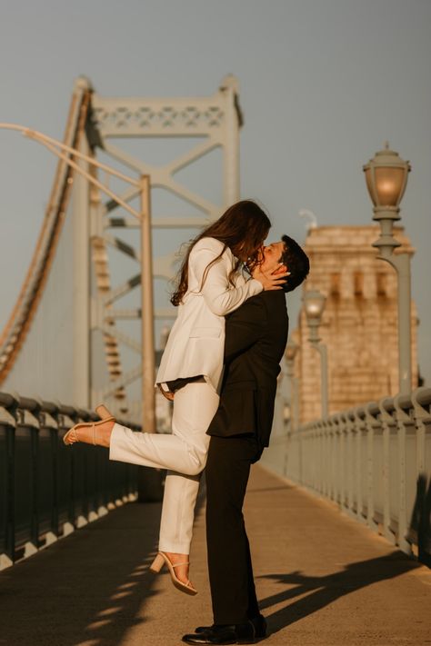 Couple poses for photo on the Ben franklin bridge in Philadelphia to celebrate their engagement. Man in black suit lifts woman in white suit up for a kiss, her foot is popped. Philadelphia Engagement Photos, Engagement Photos Couple, Classy Engagement Photos, Pictures Engagement, Photoshoot Engagement, Nyc Engagement, Vintage Photoshoot, Engagement Inspo, Classic Engagement