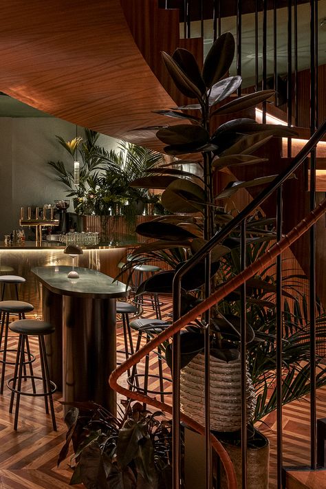 Restaurant entrance with a walnut wood spiral staircase, leading into a bar area that has marble bar tables with gold accents, light grey bar stools, and large tropical plants decorating the indoor space. Swanky Restaurant, Restaurant Ceiling, Marble Flooring, Suspended Ceiling, Wood And Marble, Vaulted Ceiling, The Restaurant, Ceiling Design, Restaurant Design