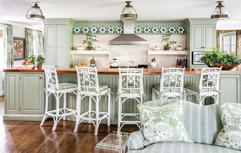 In the kitchen, a soft blue and green palette, butcher-block counters, creamy marble backsplash, and cabinetry painted “Raintree Green” by Benjamin Moore creates a country house feel. An overscale ticking stripe by Kravet on the sofa mixes with trellis wallpaper (pattern: “Fairfield” by Anna French) and a toile pattern of greenhouses, vegetables and beehives on the accent pillows and drapes (pattern: “The Allotment” by Sanderson). Interior design by James Farmer. Photo by Jeff Herr