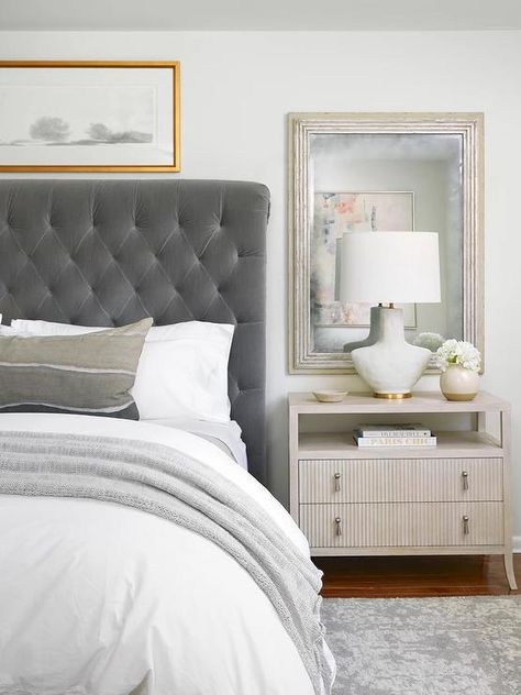 Bedroom features a dark gray velvet tufted sleigh headboard with white and gray bedding, a sliver leaf mirror mounted over a reeded nighstand lit by a white and gold lamp and a gray rug. Grey Velvet Headboard Bedroom Ideas, Gray Tufted Headboard Bedroom, Grey Tufted Bed Bedroom Ideas, Gray Velvet Bedroom Ideas, Grey Tufted Headboard Bedroom, Tufted Headboard Bedroom Decor, Dark Gray Headboard Bedroom, Dark Headboard, Grey Headboard Bedroom Ideas