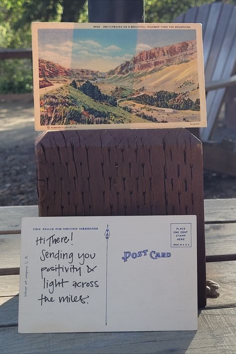 Two postcards propped up on a wooden table with a campsite in the background. A positive message is written on the back of one of the postcards.