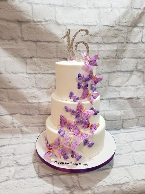 Three tiered Sweet 16 cake in all white fondant with  cascading  butterflies in different shades of purple with 16 rhinestone topper Sweet 16 Cakes With Butterflies, Sweet 15 Butterfly Cake, Sweet 16 Cake Butterfly, Purple Sweet 16 Dessert Table, Purple And White Butterfly Cake, Butterfly Themed Birthday Party Sweet 16, Purple And White Sweet 16 Cake, Quince Cakes Butterfly, Sweet 16 Cake Ideas 3 Tier