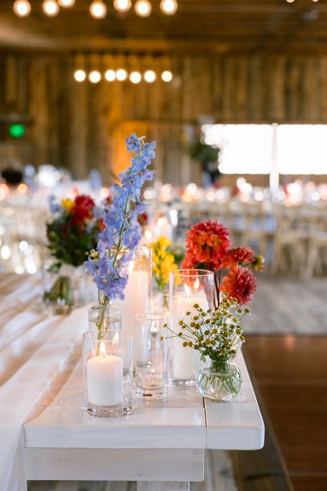 Blue delphinium, coral dahlia, yellow freeisa, feverfew chamomile bud vase and pillar candle centerpiece Wild Flower Bud Vase Centerpiece, Wildflower Bud Vase Centerpieces, Wildflower Candle Centerpiece, Floating Candles With Bud Vases, Bud Vase And Floating Candle Centerpiece, Pillar Candle And Bud Vase Centerpiece, Dahlia Bud Vase Centerpiece, Pillar Candles And Bud Vases, Delphinium Bud Vase