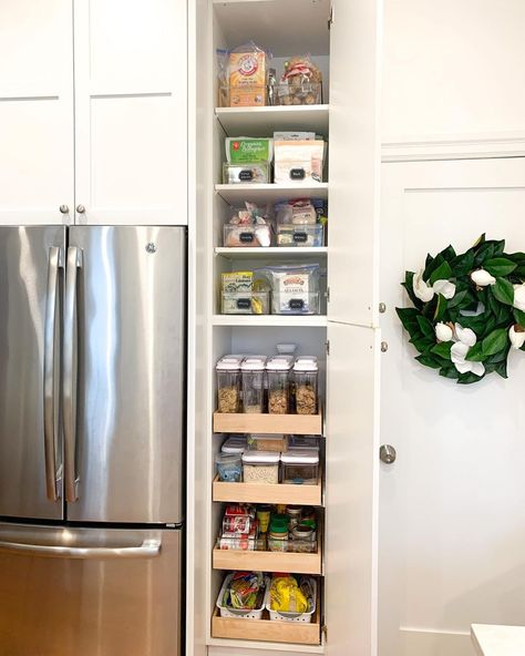 This vibrant kitchen features a large stainless steel refrigerator and bright white cabinets adorned with a leafy wreath. A narrow pantry is built into the wall and displays rows of clear bins with neat labels. Turn a small space in your kitchen into a pantry with stackable bins and canisters, and use labels to keep everything tidy. Narrow Pantry Next To Refrigerator, Fridge With Pantry Cabinet, Small Pantry For Small Kitchen, How To Add Pantry To Small Kitchen, Narrow Pantry Storage, Pantry In Open Concept Kitchen, Pantry In The Wall, Narrow Larder Cupboard, Narrow Space Next To Refrigerator