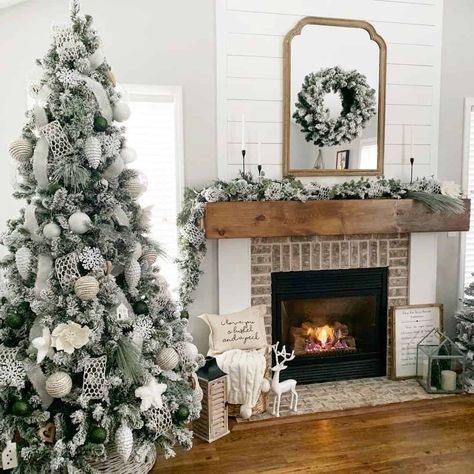 This antique fireplace possesses an exposed-brick surround, white jambs, and a large wooden mantel. A large vintage mirror with a wreath attached is mounted on the shiplap overmantel. A decorated Christmas tree stands to the left of the fireplace. Monogram Over Fireplace, Brick Fireplace Christmas Decor, Large Mirror Over Fireplace, Hamptons Christmas, White Stone Fireplaces, Large Vintage Mirror, Christmas Tree Stands, Rose Gold Christmas Tree, White Brick Fireplace