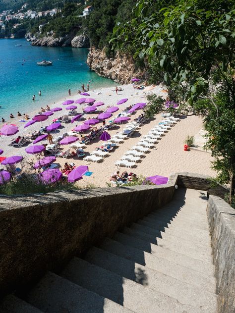 Sveti Jakov Beach in Dubrovnik Croatia, one of the best beaches in Dubrovnik. An aesthetic beach, with clear blue water, some people swimming, a few boats near the shore, purple sun umbrellas on the beach and sun loungers. The photo is taken from the top of the staircase looking down at the beautiful beach. Banje Beach Dubrovnik, Dubrovnik Aesthetic, Dubrovnik Beach, Croatia Island Hopping, Croatia Map, Viking Ocean Cruise, Lokrum Island, Plitvice National Park, Dubrovnik Old Town