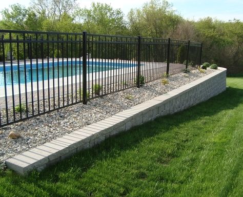 A retaining wall made of StoneWall Select block stabilizes the area around an in-ground pool. Pool Fence On Retaining Wall, Above Ground Pool Retaining Wall, Sloped Pool, Pools With Retaining Walls Sloped Backyard, Retaining Wall With Fence, Backyard Pool Fence, Pool Retaining Wall, Fence Around Pool, Landscaping Around Pool