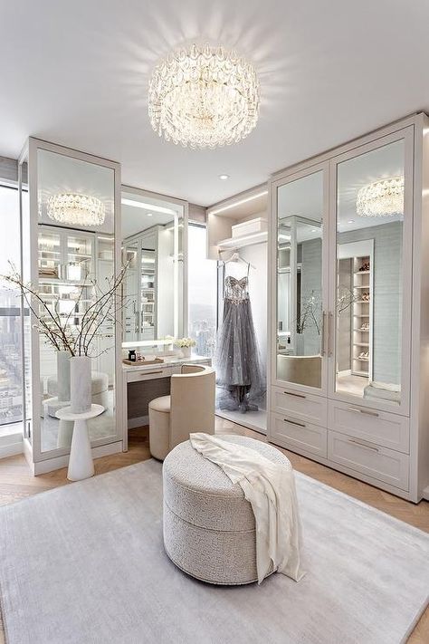 Glam light gray walk in closet illuminated by a crystal flush mount light features mirrored wardrobe cabinets, a round gray ottoman and a custom makeup vanity with cream velvet vanity chair. Gray Walk In Closet, Walk In Closet Design Ideas, Walk In Closet Vanity, Custom Makeup Vanity, Aesthetic Wardrobe Closet, Velvet Vanity Chair, Closet With Vanity, Organization Wardrobe, Gray Ottoman