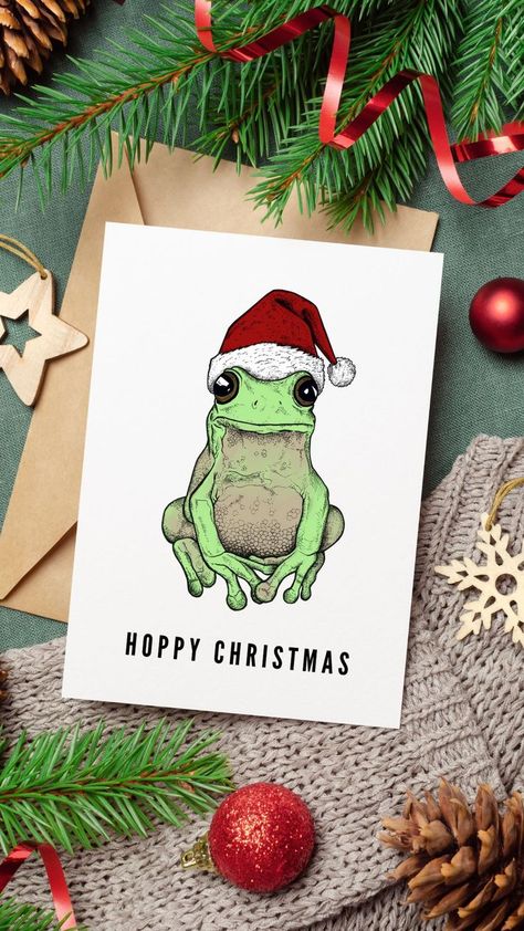 Picture shows a Christmas tableau showcasing a greeting card with a cute green frog wearing a red Santa hat and the pun "Hoppy Christmas". Christmas Frog Drawing, Christmas Frog Wallpaper, Christmas Frog Art, Frog And Toad Christmas, Frog Christmas Card, Animals With Christmas Hats, Otter Christmas Cards, Frog Christmas, Christmas Frog