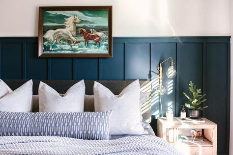 A dark wood-framed art piece is mounted above this gray upholstered bed clad in white and blue bedding. The bed is accompanied by a light wood bedside table lit by a gold and glass wall sconce. Add depth to the room with navy blue board and batten paneling behind the bed. Blue Board And Batten, White And Blue Bedding, Beige Living Room Walls, Gray Upholstered Bed, Wall Paneling Ideas, Light Wood Bed, Grey And White Bedding, Colorful Bedroom, White Wainscoting